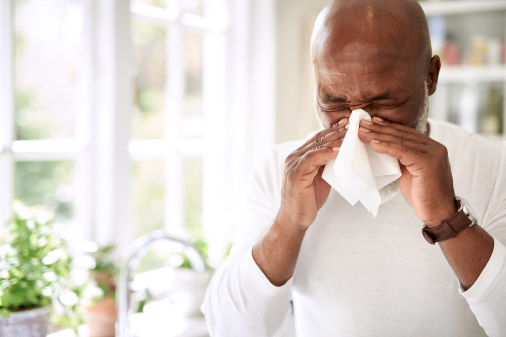 A person wearing a mask outdoors, holding tissues while surrounded by blooming flowers during allergy season.