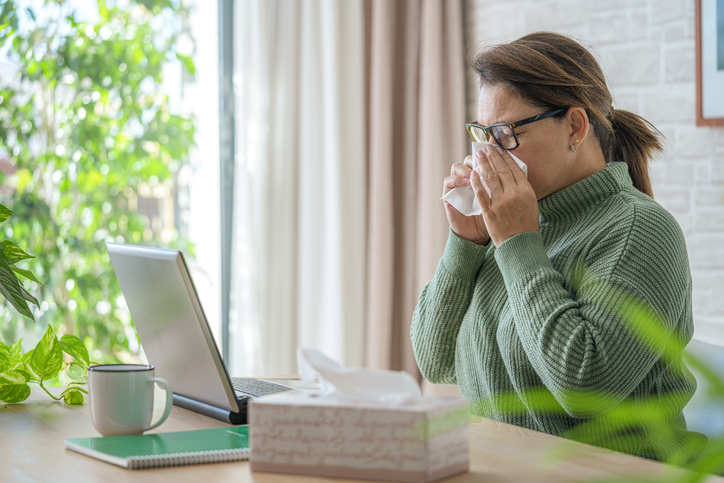 Woman with spring allergies working on laptop blowing her nose with facial tissue