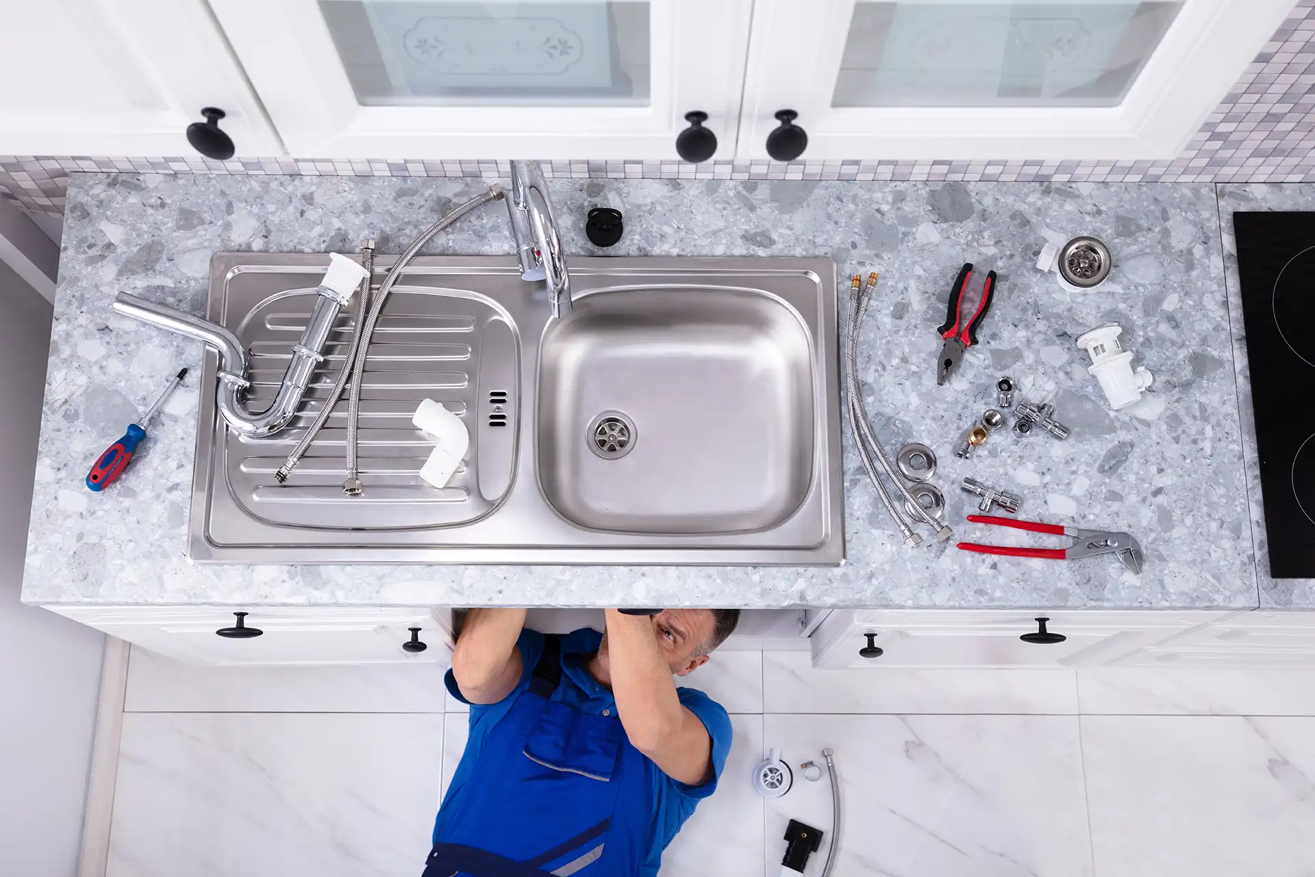 Plumber working under kitchen sink with plumbing tools laid out on countertop